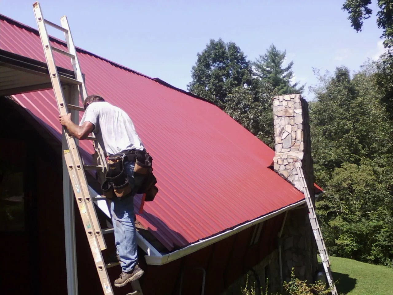 Roofer on a ladder beside a red metal roof and stone chimney — Soffit & Fascia Repair by Roofing Brothers in Farley